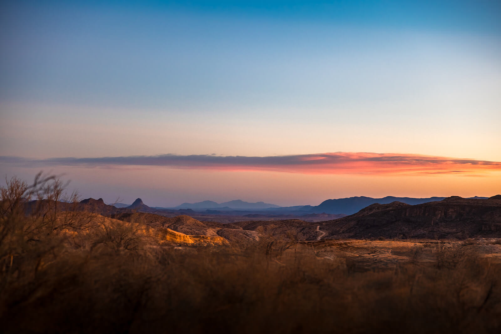 Sweeping desert landscape at dusk near Terlingua Ghost Town, Texas. Golden scrub brush fills the foreground, layered mountain ridges fade into blue haze in the distance, and a dramatic band of pink and orange clouds stretches across a gradient sky — deep amber at the horizon bleeding into soft blue overhead.